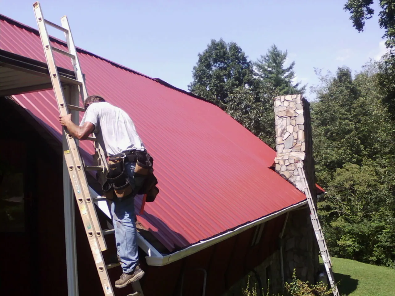 Roofer on a ladder beside a red metal roof and stone chimney — Soffit & Fascia Repair by Roofing Brothers in Tarboro
